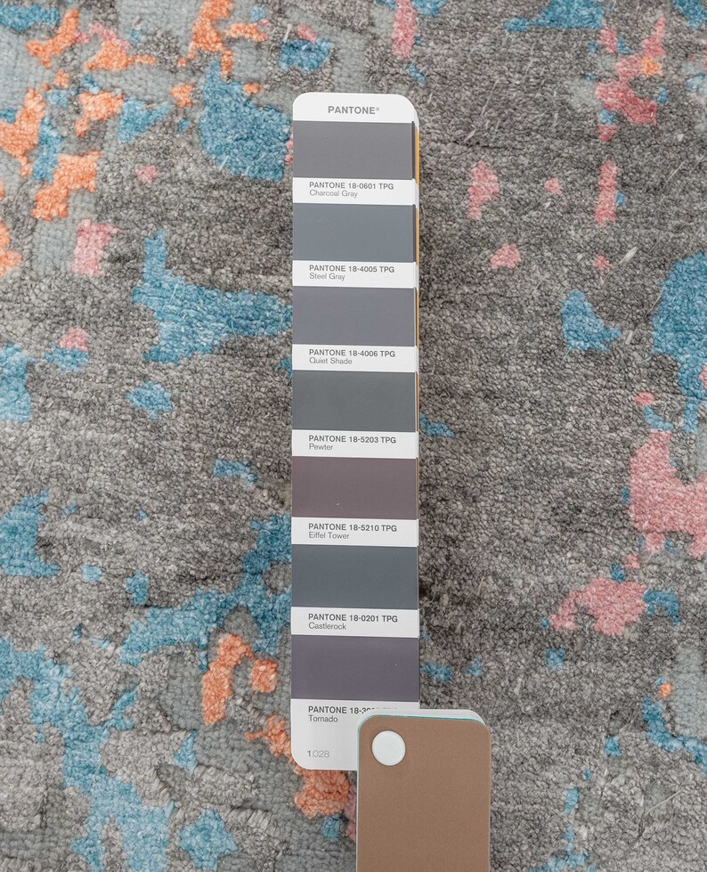 K&ouml;nnte beinhalten: Ein grauer Teppich mit abstrakten Mustern in Blau, Orange und Rosa. Rechts befindet sich eine Pantone-Farbkarte mit verschiedenen Graut&ouml;nen. Das Design des Teppichs weist ein strukturiertes, gesprenkeltes Aussehen auf.