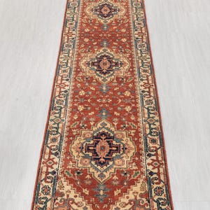 Pode incluir: Um tapete longo e retangular com um design persa tradicional. O tapete tem um fundo vermelho com uma borda de cor creme e padrões florais intrincados em tons de azul, verde e creme.