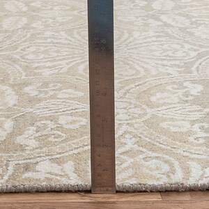 Puede incluir: Primer plano de una alfombra beige con un patr&oacute;n blanco. Una regla se coloca junto a la alfombra para mostrar el tama&ntilde;o. La regla mide 48 cm.