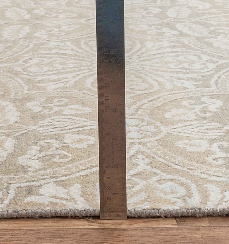 Puede incluir: Primer plano de una alfombra beige con un patr&oacute;n blanco. Una regla se coloca junto a la alfombra para mostrar el tama&ntilde;o. La regla mide 48 cm.