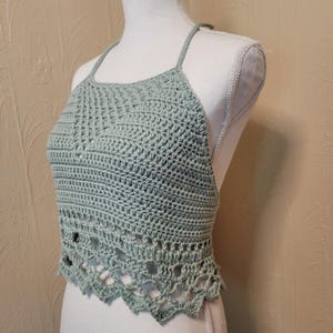 May include: A light green, handmade crochet halter top. The top features a textured stitch pattern and a scalloped edge. The halter straps are thin and the top is displayed on a mannequin.