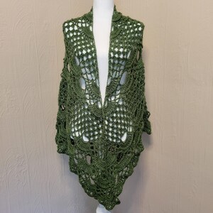 May include: A green crocheted shawl with a delicate lace pattern. The shawl is draped over a mannequin and has a triangular shape.
