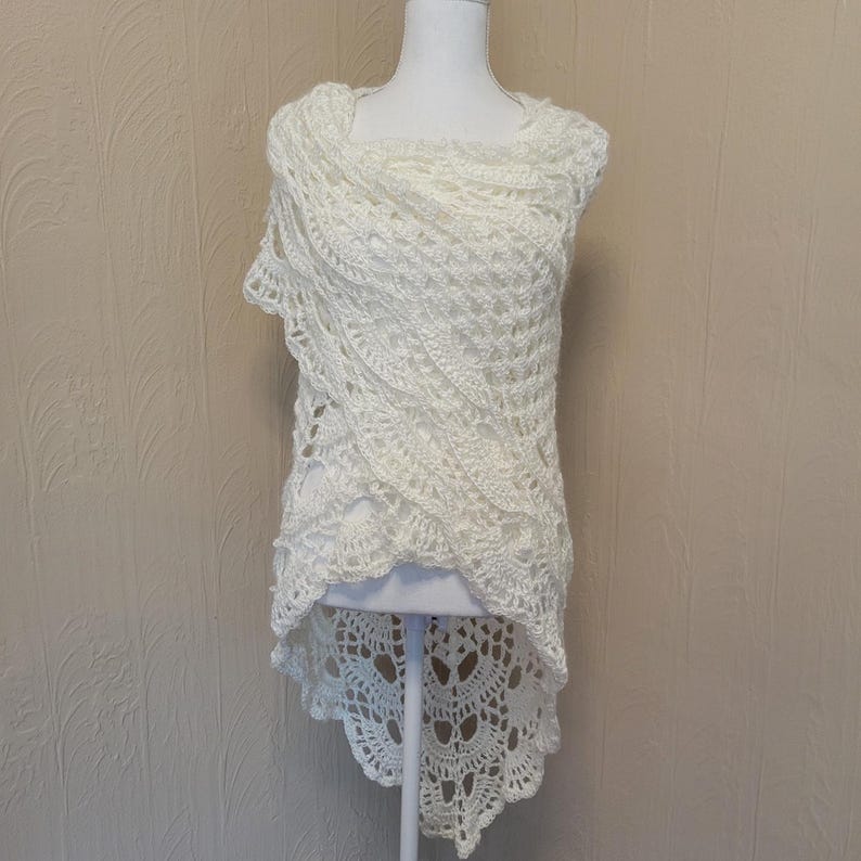 May include: White crocheted shawl with a high-low hemline. The shawl is made with a delicate openwork pattern and has a scalloped edge.