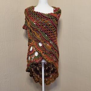 May include: A crochet shawl featuring an intricate, openwork pattern in a variegated color palette of deep forest green, burnt orange, mustard yellow, and dark burgundy. The garment has a high-low hemline with a scalloped edge and draped design.