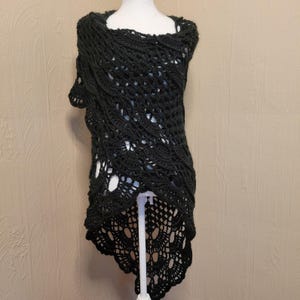 Puede incluir: Un chal negro de ganchillo con un diseño de tejido abierto, exhibido en un maniquí. El chal presenta un escote cuadrado y un diseño asimétrico. El patrón de ganchillo incluye espacios abiertos, creando una apariencia delicada y aireada. El chal es de color negro liso.