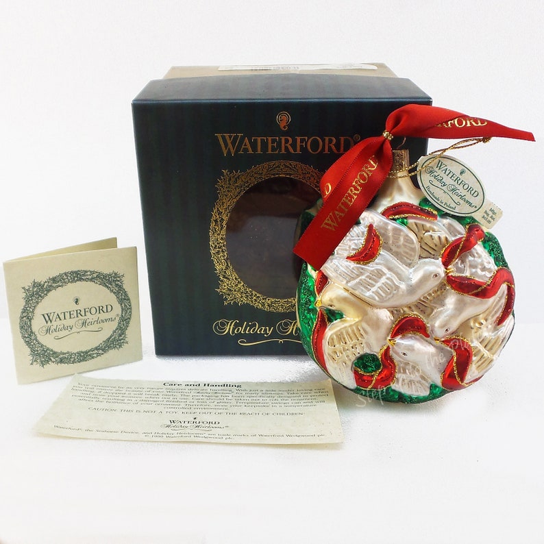 May include: A Waterford Holiday Heirloom glass ornament featuring two white doves with red ribbons, surrounded by green leaves and a gold background. The ornament is in its original box with a tag that reads "Waterford Holiday Heirloom".