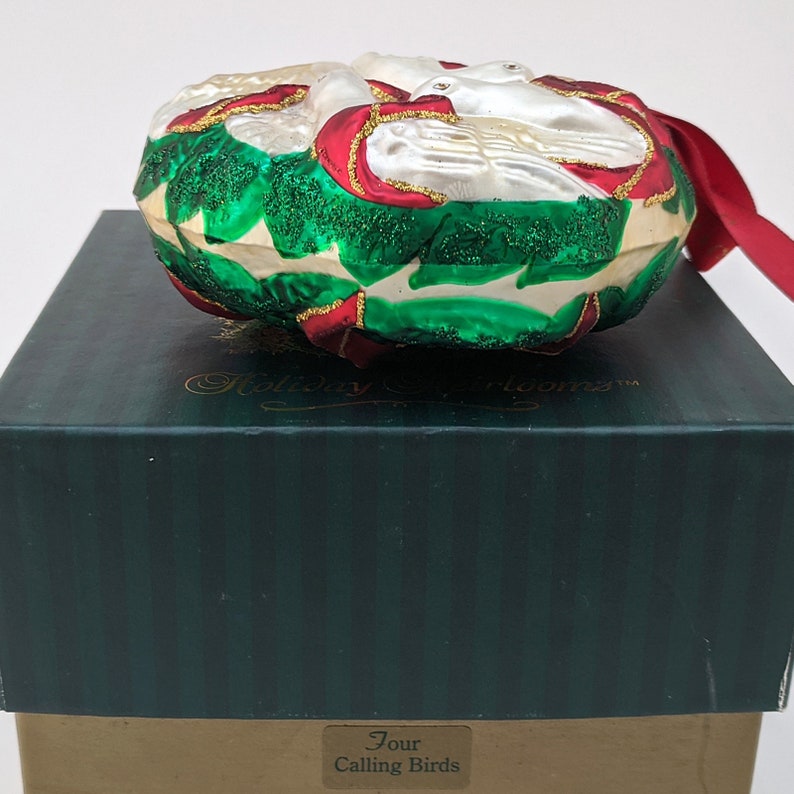 May include: A glass Christmas ornament shaped like a wreath with two white doves inside. The wreath is decorated with red and gold accents on a green background. The ornament is in a box with the words "Four Calling Birds" on the label.