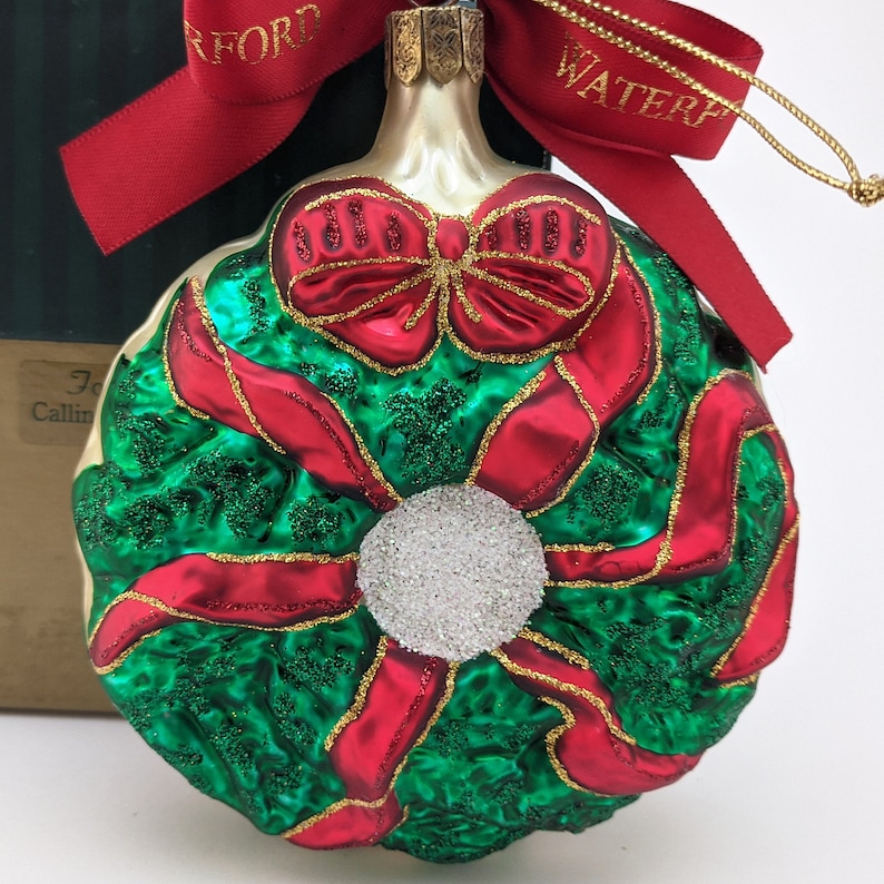 May include: A glass Christmas ornament shaped like a wreath with red ribbon and a white center. The ornament has a gold glitter finish.