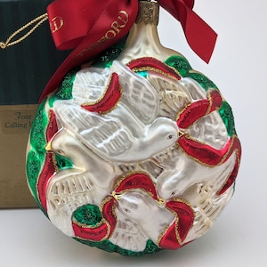 May include: A glass Christmas ornament featuring four white doves with red and green accents. The doves are depicted in a circular pattern, with their wings spread out. The ornament is decorated with gold glitter.