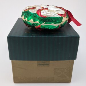 May include: A white glass Christmas ornament with a green and red holly design and two white doves. The ornament is in a green and brown box with the text "Four Calling Birds".