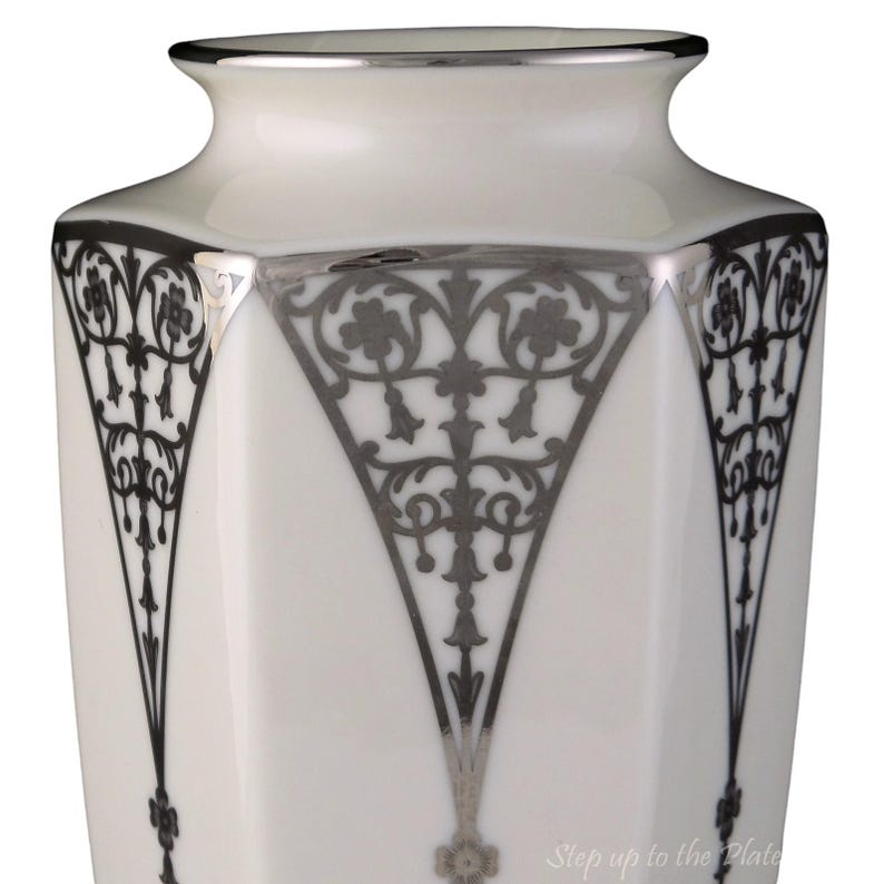 Puede incluir: Jarr&oacute;n de porcelana blanca con un dise&ntilde;o geom&eacute;trico plateado. El jarr&oacute;n tiene una forma hexagonal y un borde plateado.
