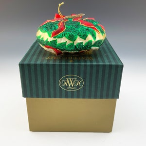 May include: A Waterford Christmas ornament in the shape of a poinsettia with green leaves and red petals. The ornament is in its original box, which is green with white stripes and the Waterford logo.
