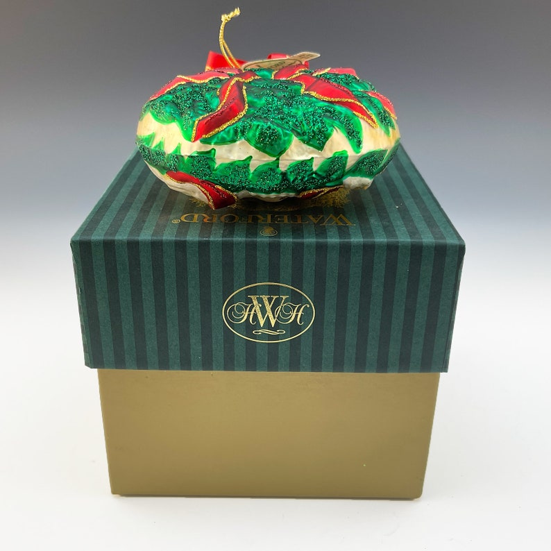May include: A Waterford Christmas ornament in the shape of a poinsettia with green leaves and red petals. The ornament is in its original box, which is green with white stripes and the Waterford logo.
