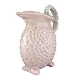 May include: A pink ceramic pitcher with a textured surface and a white handle. The pitcher has a rounded body and three feet.