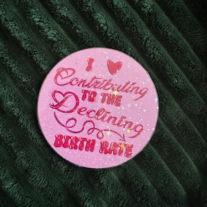 May include: A pink, round sticker with the text "I ❤️ Contributing to the Declining Birth Rate" in a cursive font. The sticker has a glittery, holographic effect and is set against a dark green, textured background.