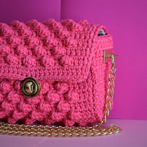 K&ouml;nnte beinhalten: Eine pinkfarbene geh&auml;kelte Handtasche mit einem strukturierten, blasenartigen Muster. Die Tasche hat einen goldfarbenen Kettenriemen und einen Verschluss. Der Hintergrund ist ein leuchtendes Pink, das die Farbe der Tasche erg&auml;nzt.