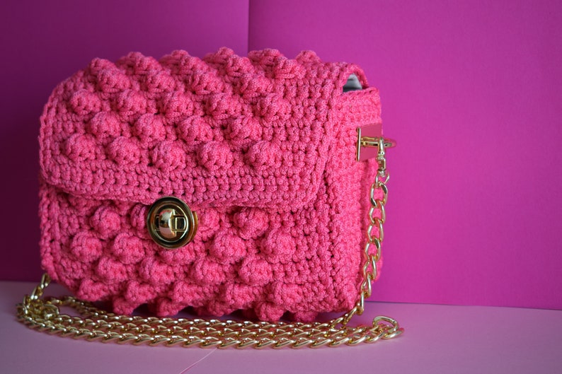 K&ouml;nnte beinhalten: Eine pinkfarbene geh&auml;kelte Handtasche mit einem strukturierten, blasenartigen Muster. Die Tasche hat einen goldfarbenen Kettenriemen und einen Verschluss. Der Hintergrund ist ein leuchtendes Pink, das die Farbe der Tasche erg&auml;nzt.