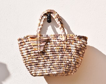 Könnte beinhalten: Eine gewebte braune, weiße und beige Tragetasche mit einem braunen Ledergriff. Die Tasche hat ein einzigartiges gewebtes Design und einen kleinen braunen Lederfleck auf der Vorderseite.