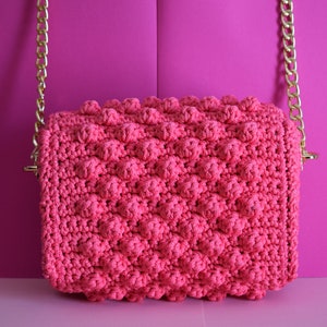 K&ouml;nnte beinhalten: Eine pinkfarbene geh&auml;kelte Handtasche mit einem strukturierten Noppenmuster. Die Tasche hat eine rechteckige Form und einen goldfarbenen Kettenriemen. Der Hintergrund ist einfarbig pink.