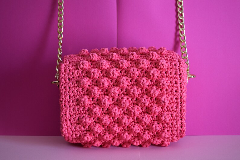 K&ouml;nnte beinhalten: Eine pinkfarbene geh&auml;kelte Handtasche mit einem strukturierten Noppenmuster. Die Tasche hat eine rechteckige Form und einen goldfarbenen Kettenriemen. Der Hintergrund ist einfarbig pink.