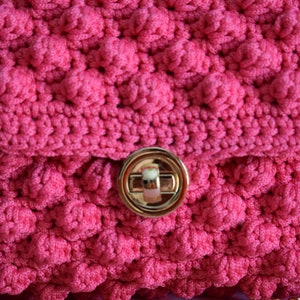 K&ouml;nnte beinhalten: Nahaufnahme einer handgefertigten, leuchtend rosa H&auml;keltasche mit einem strukturierten, erhabenen Blasenstichmuster. Die Tasche hat einen goldfarbenen Verschluss und einen Kettenriemen. Der Klappenverschluss wird durch einen goldfarbenen Drehverschluss gesichert.