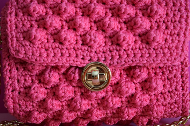 K&ouml;nnte beinhalten: Nahaufnahme einer handgefertigten, leuchtend rosa H&auml;keltasche mit einem strukturierten, erhabenen Blasenstichmuster. Die Tasche hat einen goldfarbenen Verschluss und einen Kettenriemen. Der Klappenverschluss wird durch einen goldfarbenen Drehverschluss gesichert.