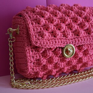 K&ouml;nnte beinhalten: Eine pinkfarbene geh&auml;kelte Handtasche mit einem strukturierten, erhabenen Muster. Die Tasche hat einen goldfarbenen Kettenriemen und einen goldfarbenen Verschluss. Die Tasche steht vor einem pinkfarbenen Hintergrund.
