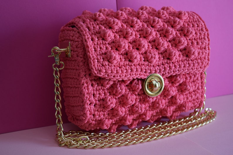 K&ouml;nnte beinhalten: Eine pinkfarbene geh&auml;kelte Handtasche mit einem strukturierten, erhabenen Muster. Die Tasche hat einen goldfarbenen Kettenriemen und einen goldfarbenen Verschluss. Die Tasche steht vor einem pinkfarbenen Hintergrund.