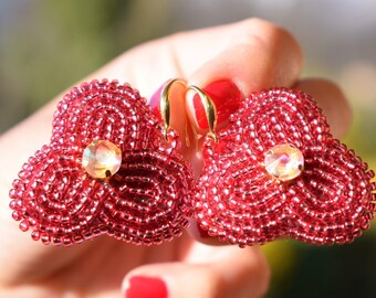 Könnte beinhalten: Ein Paar rote Perlenohrringe mit einem Kristall in der Mitte. Die Ohrringe sind in Form einer Blume mit drei Blütenblättern.