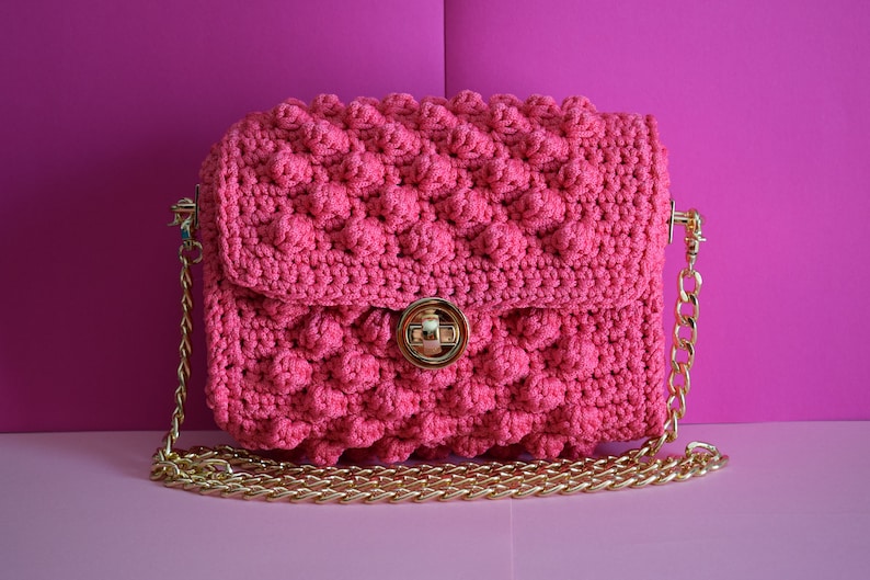 K&ouml;nnte beinhalten: Eine pinkfarbene H&auml;keltasche mit einem strukturierten, blasenartigen Muster. Die Tasche hat einen goldfarbenen Kettenriemen und einen goldenen Verschluss. Der Hintergrund ist ein Farbverlauf in Rosat&ouml;nen.