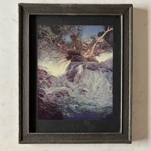 May include: Framed art print depicting a nude figure seated on a rock formation near a waterfall. The artwork features a dark, textured frame and a black matting. The color palette includes blues, greens, and browns, creating a serene and naturalistic scene.