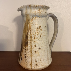 May include: A handmade ceramic pitcher with a cream-colored base and a brown and tan speckled design. The pitcher has a curved handle and a spout for pouring. The pitcher is approximately 8 inches tall.
