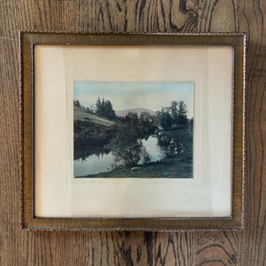 May include: A framed landscape print featuring a river scene with trees and a distant mountain. The print is set within a gold-toned, textured frame. The artwork depicts a serene natural environment with soft colors.