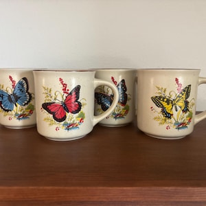 Könnte beinhalten: Ein Set aus vier cremefarbenen Keramikbechern mit gesprenkelter Oberfläche. Jeder Becher zeigt eine farbenfrohe Schmetterlingsillustration, darunter blaue, rote und gelbe Arten, umgeben von grünem Blattwerk und kleinen roten Beeren.