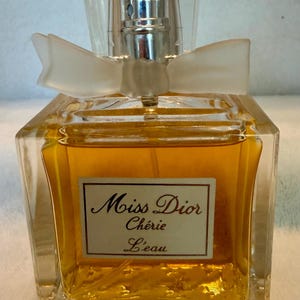 May include: A square glass perfume bottle filled with amber liquid, featuring a clear cap and a translucent bow at the neck. A white rectangular label on the front displays the text Miss Dior Chérie L'eau in dark brown script and serif lettering.