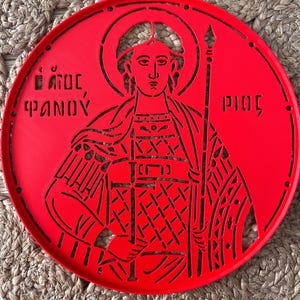 Puede incluir: Una bandeja decorativa circular de color rojo brillante con un diseño recortado de una figura religiosa que sostiene un báculo. La figura tiene un halo y está rodeada de texto griego. La bandeja descansa sobre un salvamanteles beige tejido.