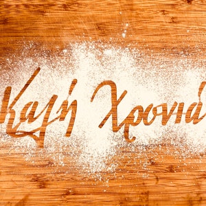 May include: The words "Kalí Khronía!" are written in white flour on a brown wooden surface.