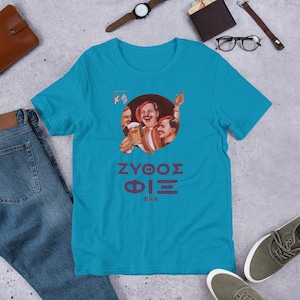 Puede incluir: Una camiseta azul turquesa con una imagen de estilo vintage de tres hombres sosteniendo una jarra de cerveza. La imagen está rodeada por un círculo y el texto "ZYOOΣ ΦΙΞ 1864" está impreso debajo de la imagen.