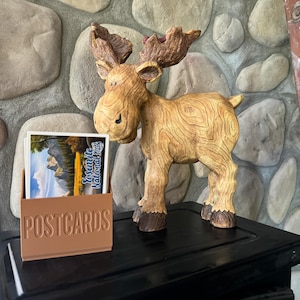 May include: A wooden moose figurine with a brown and tan wood grain finish stands on a black surface. A brown wooden postcard holder with the word "POSTCARDS" embossed on the front holds a postcard with a picture of Yosemite National Park.