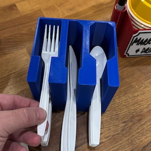May include: A blue plastic utensil holder with three compartments, holding a white plastic fork, knife, and spoon. The holder is rectangular and has a textured surface. The utensils are arranged vertically.