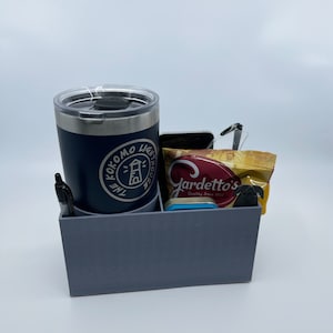 May include: A gray desk organizer with four compartments. The organizer holds a blue tumbler with a white logo that says "The Kokomo Lighthouse", a black pen, a cell phone, and a bag of Gardetto's snacks.