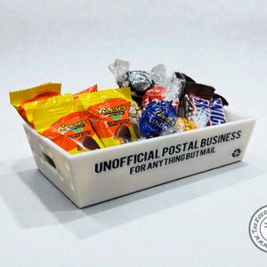 May include: A white rectangular basket filled with assorted wrapped candies, including Reese's peanut butter cups and Snickers bars. The basket has cut-out handles and text that reads "UNOFFICIAL POSTAL BUSINESS FOR ANYTHING BUT MAIL."