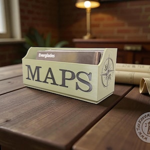 May include: A light green wooden map holder with the word "MAPS" in dark gray lettering. A compass rose design is on the side. The holder contains maps labeled "Everglades".