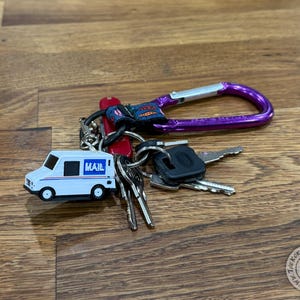 May include: A keychain with keys, a purple carabiner, and a white miniature mail truck charm with the word "MAIL" on the side. The keys are attached to a silver keyring.