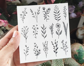 May include: A square sheet of paper featuring a collection of delicate, black line-drawn floral and botanical illustrations. The designs include various flowers, leaves, and sprigs, perfect for temporary tattoos or craft projects.