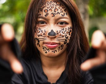 Puede incluir: Una persona con maquillaje de leopardo en la cara, incluyendo manchas en la frente y mejillas, una nariz negra y bigotes. La persona lleva una chaqueta con capucha negra. El fondo es follaje verde borroso.