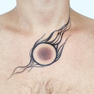 Peut inclure: Un tatouage abstrait noir et blanc sur la poitrine d'une personne. Le motif présente un cercle avec une tache sombre au centre et une série de lignes ondulées s'étendant du cercle.