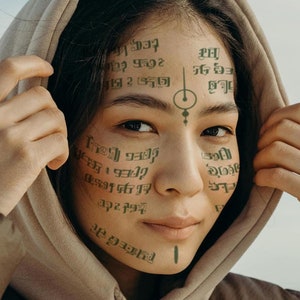 May include: A person wearing a brown hooded sweatshirt with green text and symbols covering their face. The text is in a foreign language.