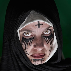May include: A person wearing a black hooded garment with a white wimple. Black makeup streaks down from the eyes and mouth. A black cross is painted on the forehead. The background is a dark green.