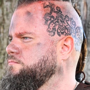 Puede incluir: Vista de perfil de una persona con barba espesa y oscura y un lado de la cabeza rapado con un tatuaje negro de nudos intrincados. La piel muestra manchas de maquillaje oscuro y la persona viste una prenda de cuero marrón con cordones.